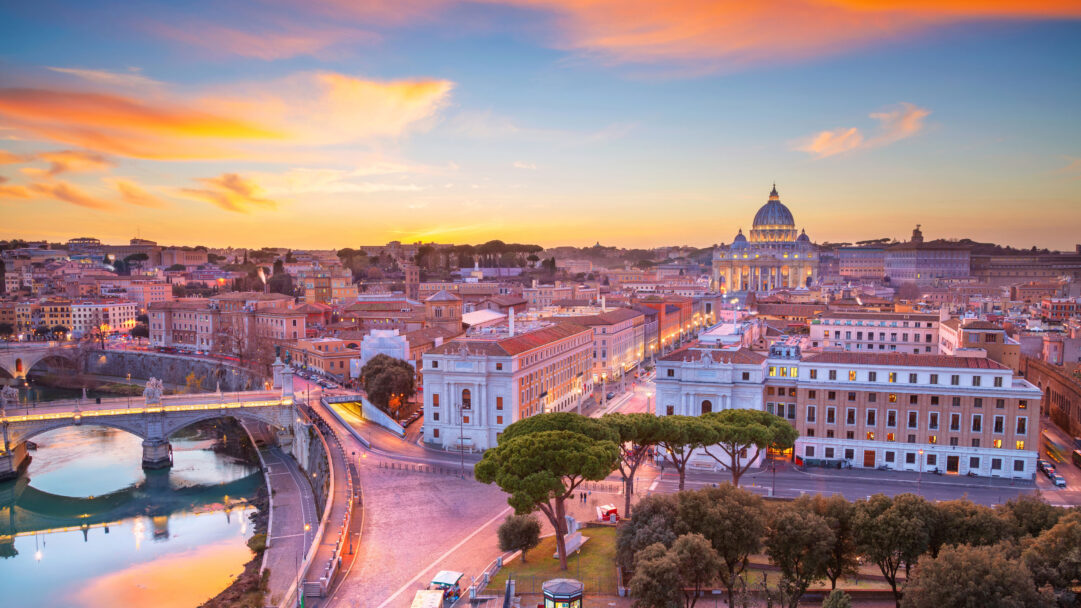 A glorious 4K wallpaper capturing the expansive sunset skyline of Rome, prominently featuring St. Peter's Basilica and the Vatican City bathed in twilight. The sky explodes with vibrant oranges and pinks, contrasting beautifully with the deepening blues, as the city lights begin to glow, creating a serene and majestic evening atmosphere.