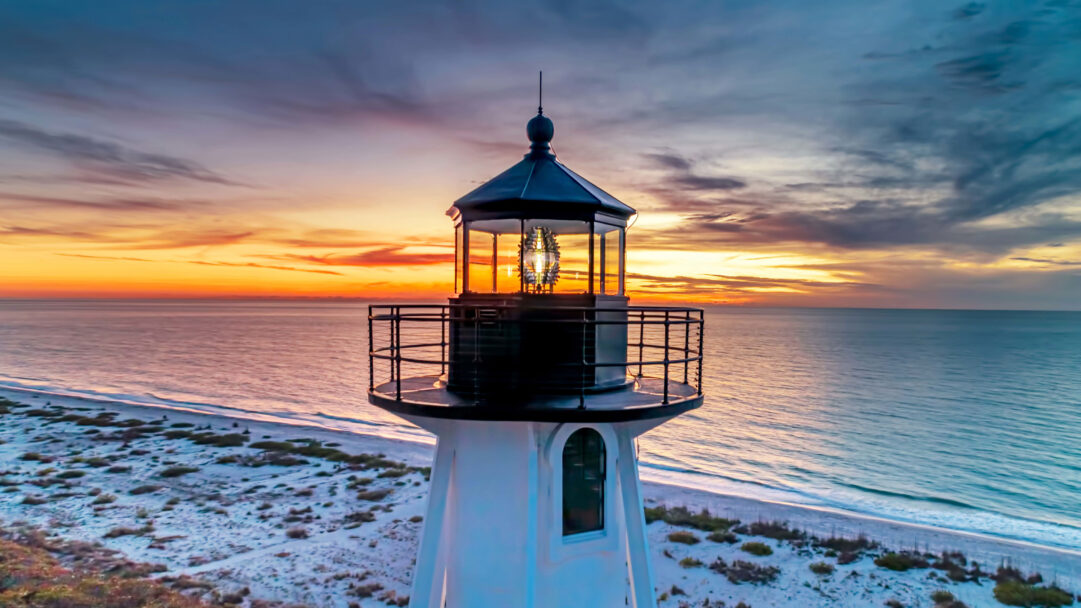 A vibrant 4K wallpaper showcasing the Gasparilla Island Rear Range Light standing prominently on a white sand beach at sunset. The scene is bathed in the dramatic hues of the vibrant sunset, with fiery oranges and deep blues reflecting across the tranquil ocean and highlighting the lighthouse's beacon.