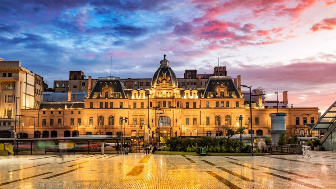 An atmospheric 4K wallpaper displays the majestic Buenos Aires Constitución Station, brightly lit at dusk. The vibrant sunset sky, painted with hues of purple, orange, and blue, dramatically contrasts with the station's warm glow and the shimmering reflections on the wet plaza below.