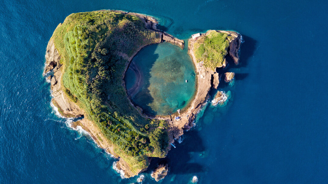 A breathtaking 4K wallpaper showcases an aerial view of the crescent-shaped Vila Franca Islet off São Miguel Island. Its vibrant natural pool, enclosed by the green-covered volcanic rock, glows with turquoise waters that contrast sharply with the surrounding deep blue Atlantic Ocean.