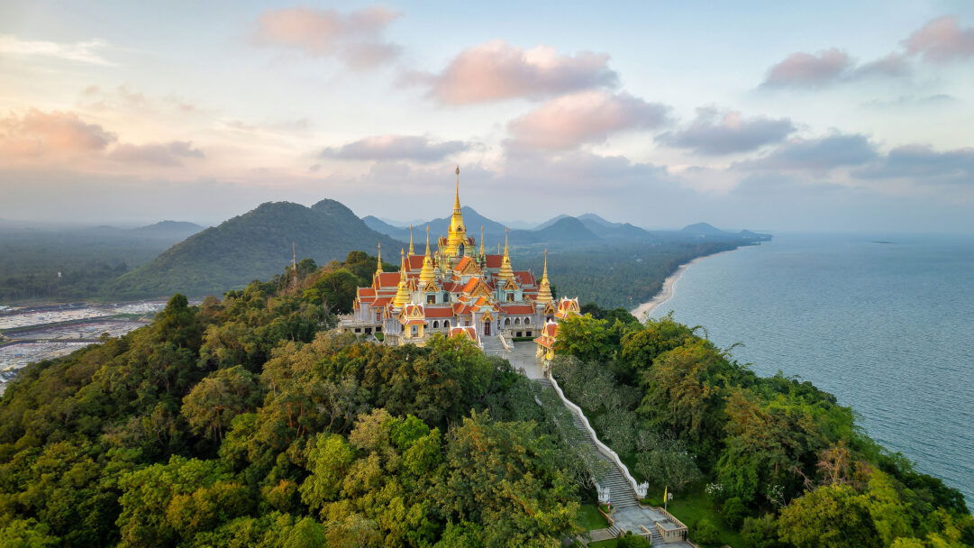 A magnificent 4K wallpaper presents the ornate Wat Tang Sai Temple grandly situated on a forested hill, overlooking the pristine Ban Krut coastline and tranquil sea. The temple's golden spires glow warmly under the soft, pastel hues of a coastal sunset, creating a serene and breathtaking landscape.