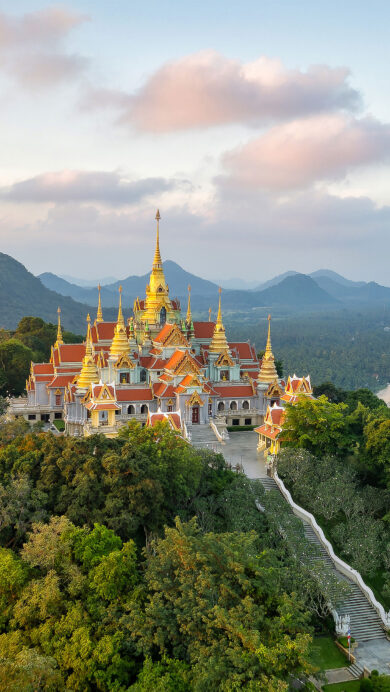 A magnificent 4K wallpaper presents the ornate Wat Tang Sai Temple grandly situated on a forested hill, overlooking the pristine Ban Krut coastline and tranquil sea. The temple's golden spires glow warmly under the soft, pastel hues of a coastal sunset, creating a serene and breathtaking landscape.