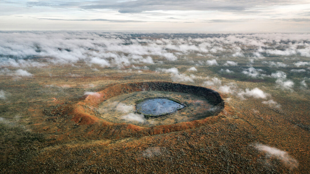 An awe-inspiring 4K wallpaper presents an aerial view of the majestic Wolfe Creek Crater nestled within the expansive Australian desert landscape. Its sharply defined, reddish-orange rim encircles a tranquil dark blue pool, with wisps of low-hanging clouds drifting across the textured, scrub-dotted terrain, enhancing the remote and grand scale.