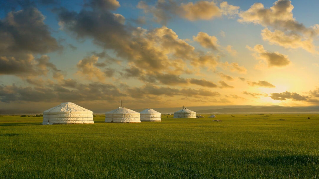 An immersive 4K wallpaper featuring several traditional white yurts scattered across the vast, lush Mongolian grasslands. The scene is bathed in the warm, golden light of a spectacular sunset, creating a serene mood as the sky transitions from soft blues to brilliant oranges above distant hills.