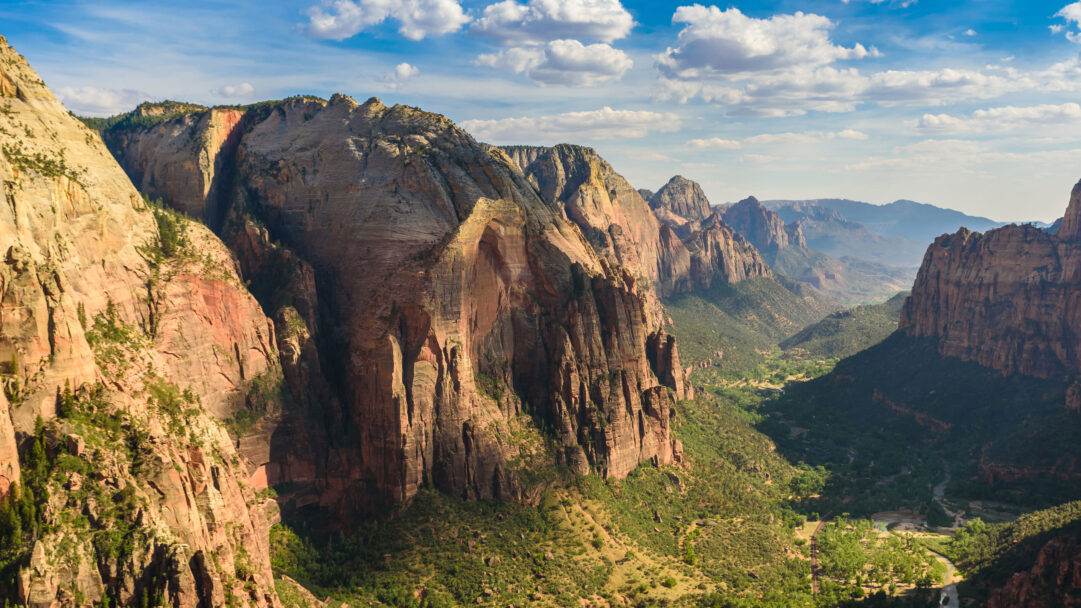 An immersive 4K wallpaper presents the majestic landscape of Zion National Park in Utah, showcasing a deep valley carved between towering, multi-hued sandstone cliffs. Golden sunlight illuminates the upper sections of the colossal rock formations, casting dramatic shadows into the verdant, tree-lined canyon floor where a river gently meanders.