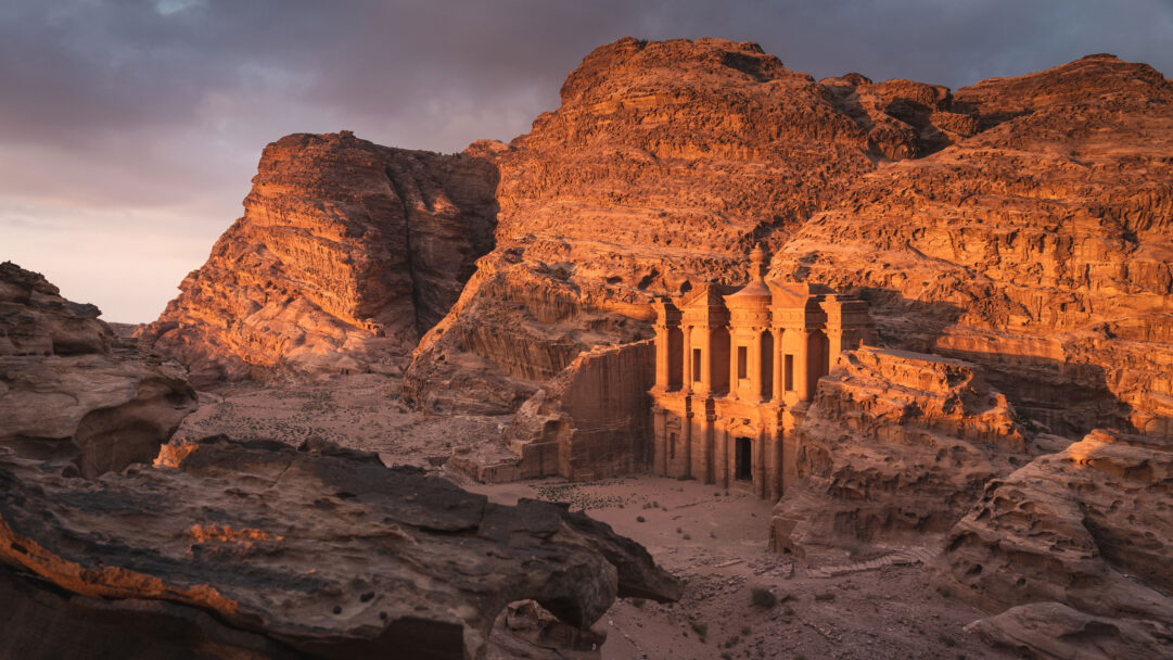 A majestic 4K wallpaper presents the ancient Ad Deir Monastery, carved directly into the towering sandstone cliffs of Petra, Jordan. Golden sunlight bathes the ornate facade and surrounding rugged landscape in a breathtaking display of warm orange and red tones under a dramatic sky.