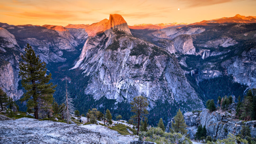 An inspiring 4K wallpaper showcasing Half Dome illuminated by alpenglow in the vast landscape of Yosemite National Park. The iconic granite face of Half Dome glows a brilliant orange-red against a twilight sky, creating a breathtaking contrast with the shadowed, rugged mountains and the serene, deep blue valleys below.