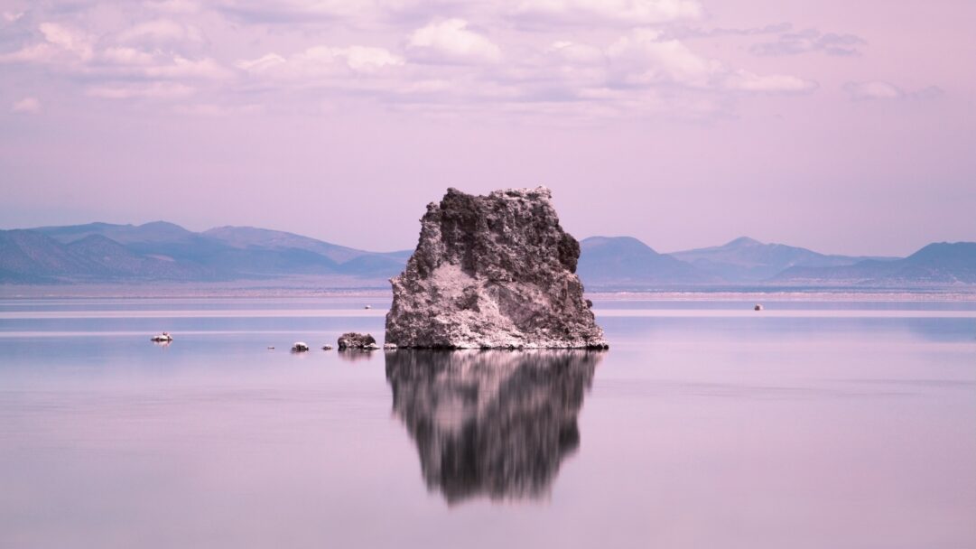 A stunning 4K wallpaper showcases a rugged beach rock formation standing prominently in a calm lake, with distant mountains under a tranquil pink sky. The rock's perfect reflection shimmers on the water's surface, mirroring the soft lavender hues of the serene sky and creating a dreamlike, ethereal mood.