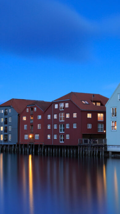 An enchanting 4K wallpaper presents a row of colorful waterfront houses in Trondheim, Norway, during the blue hour twilight. Their warm, glowing windows cast long, vibrant reflections across the smooth, deep blue water, creating a peaceful and inviting evening scene.