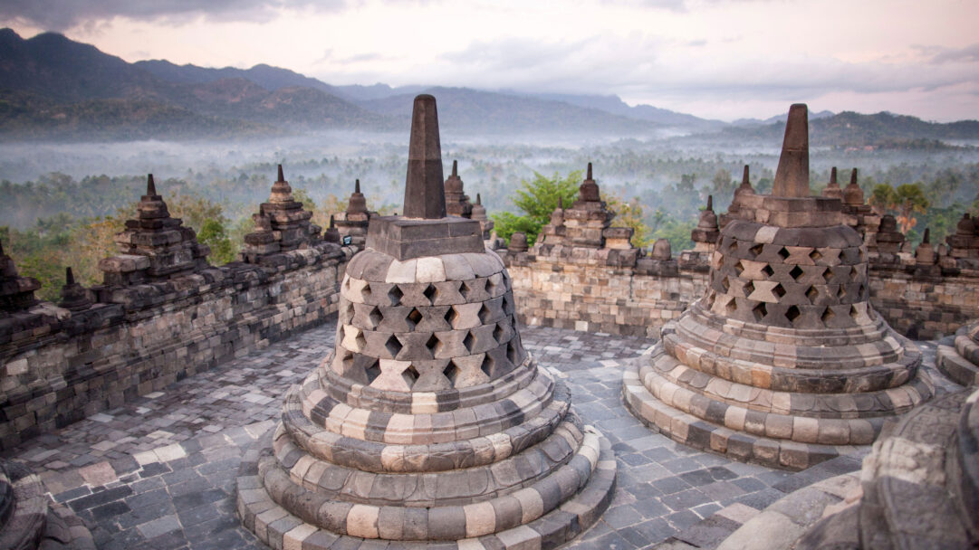 A serene 4K wallpaper featuring the ancient Borobudur Temple stupas on Java, Indonesia, overlooking a vast, misty landscape. Intricate grey stone stupas stand prominently against a soft, overcast sky, while a thick mist blankets the distant green mountains and valleys, evoking a profound sense of peace.
