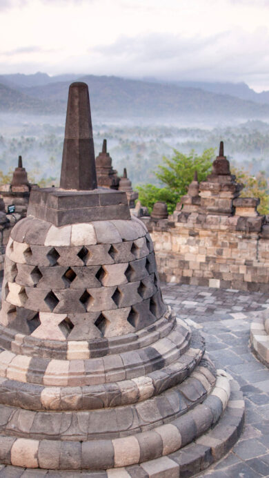 A serene 4K wallpaper featuring the ancient Borobudur Temple stupas on Java, Indonesia, overlooking a vast, misty landscape. Intricate grey stone stupas stand prominently against a soft, overcast sky, while a thick mist blankets the distant green mountains and valleys, evoking a profound sense of peace.