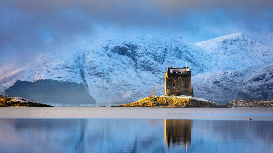 A serene 4K wallpaper depicts Castle Stalker, a historic stone castle perched on a small, sunlit island within Loch Laich, Scotland, set against a backdrop of majestic snow-covered mountains in winter. The tranquil waters of the loch flawlessly mirror the castle and the cool blue sky, emphasizing the stark beauty and calm of the Scottish winter landscape.