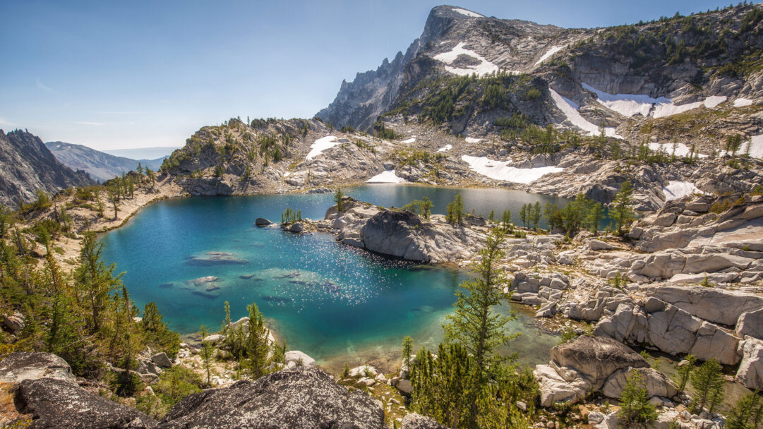 An enchanting 4K wallpaper portrays the pristine Crystal Lake nestled within the rugged Alpine Lakes Wilderness mountain landscape. Its crystal-clear turquoise waters glitter with sunlit sparkles, surrounded by majestic granite peaks dotted with snow patches and resilient evergreen trees under a clear blue sky.
