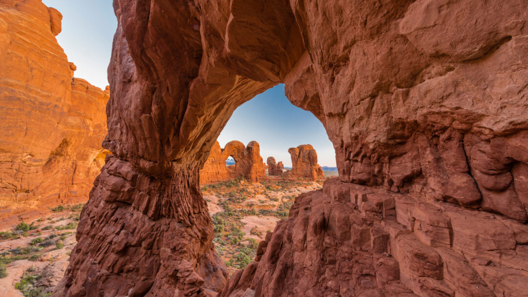 A breathtaking 4K wallpaper showcasing Double Arch perfectly framed through the vast opening of Cove Arch in a majestic desert landscape. The golden hour light illuminates the intricate textures of the towering red sandstone, highlighting the dramatic natural architecture against a soft blue sky.