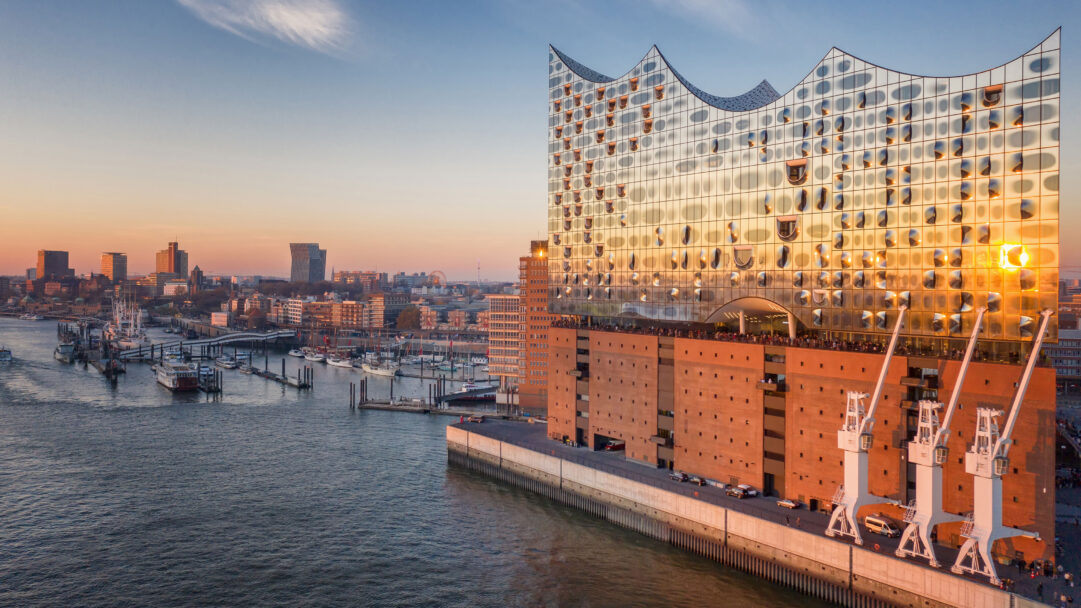 A spectacular 4K wallpaper captures the striking Elbphilharmonie Concert Hall at sunset on the bustling Hamburg waterfront. Golden sunlight brilliantly reflects off the building's distinctive wavy glass facade, illuminating the surrounding cityscape and calm waters of the harbor.