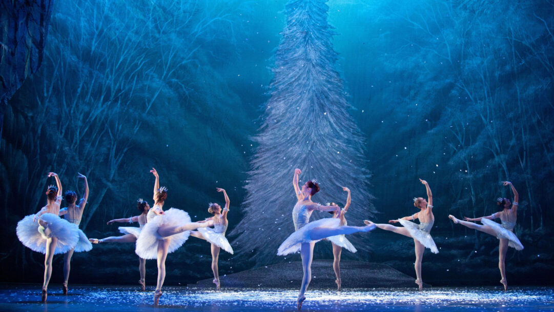 An enchanting 4K wallpaper capturing the English National Ballet performing 'The Nutcracker' on a stage transformed into a magical winter forest. The scene features graceful ballerinas in white tutus, bathed in cool blue light, with shimmering snowflakes falling around a tall, frosted pine tree.