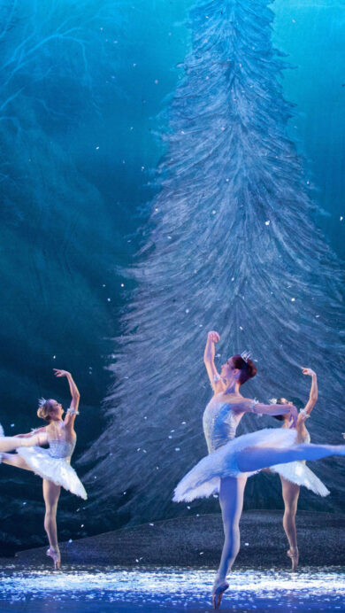 An enchanting 4K wallpaper capturing the English National Ballet performing 'The Nutcracker' on a stage transformed into a magical winter forest. The scene features graceful ballerinas in white tutus, bathed in cool blue light, with shimmering snowflakes falling around a tall, frosted pine tree.