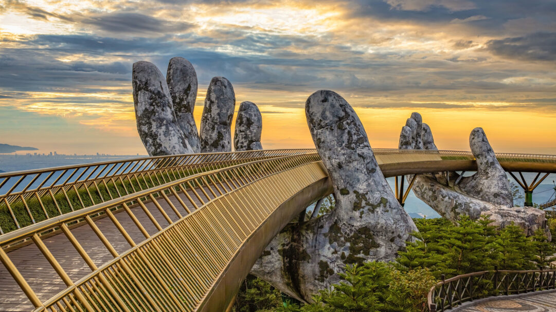 An awe-inspiring 4K wallpaper presents the iconic Golden Bridge at Bà Nà Hills, Da Nang, Vietnam, gracefully held aloft by giant moss-covered stone hands. The dramatic sunset bathes the scene in warm golden and soft grey light, highlighting the bridge's gleaming railings and the distant Da Nang city skyline.