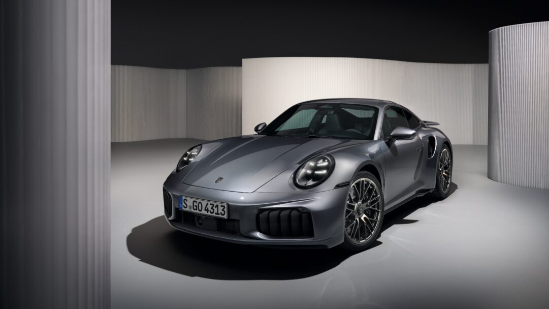 A sleek 4K wallpaper presenting a grey Porsche 911 Turbo S sports car parked diagonally in a minimalist, modern studio. The car's polished grey body gleams under precise studio lighting, creating sharp reflections and deep shadows against the subtly textured white and dark grey background.