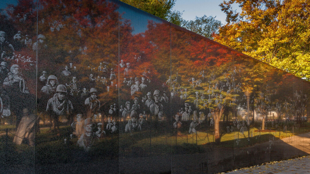 A poignant 4K wallpaper featuring the Korean War Veterans Memorial Mural Wall in Washington D.C., etched with the solemn faces of soldiers. The polished black granite surface vividly reflects the vibrant red, orange, and gold hues of the surrounding autumn trees, blending commemoration with seasonal beauty.