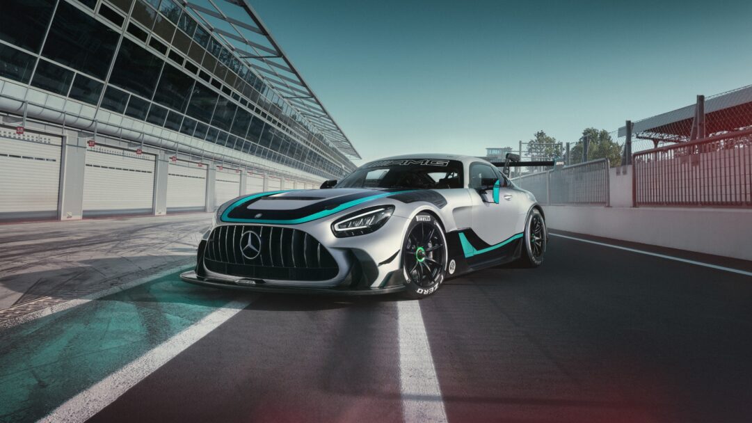 A captivating 4K wallpaper presenting the Mercedes-AMG GT2 Edition W16 race car parked on the asphalt of a sunlit racing track, with pit garages and a modern building in the background. Its sleek silver body, accentuated by vibrant teal and black racing stripes, glistens under the bright sky, highlighting the vehicle's formidable design and powerful stance.