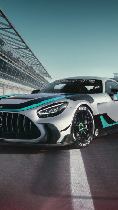 A captivating 4K wallpaper presenting the Mercedes-AMG GT2 Edition W16 race car parked on the asphalt of a sunlit racing track, with pit garages and a modern building in the background. Its sleek silver body, accentuated by vibrant teal and black racing stripes, glistens under the bright sky, highlighting the vehicle's formidable design and powerful stance.