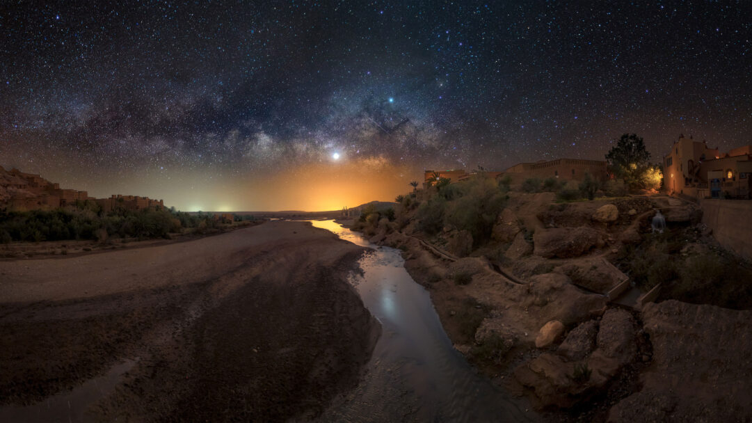 An awe-inspiring 4K wallpaper depicting the magnificent Milky Way Galaxy arching over the historic fortified village of Aït Benhaddou in Morocco at night. The brilliant cosmic dust and countless stars of the galaxy stretch across the dark sky, dramatically contrasting with the warm, amber glow from the ancient earthen structures that line the winding river below.
