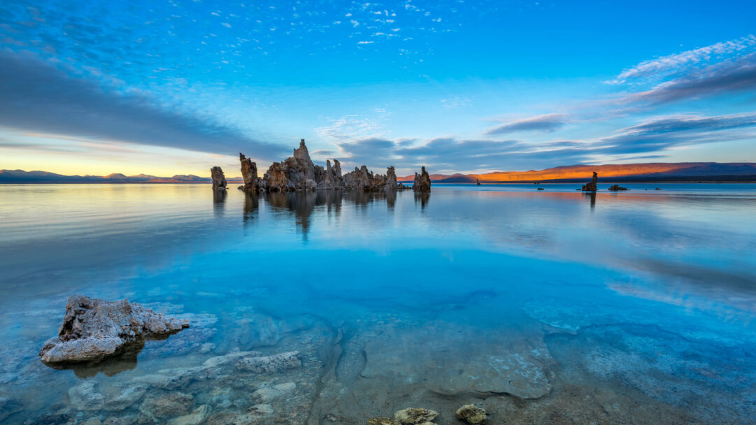 An awe-inspiring 4K wallpaper featuring the striking tufa formations of Mono Lake, rising from the tranquil waters under a vivid sunset sky. The calm, reflective surface of the lake perfectly mirrors the dramatic clouds and the warm golden glow of the setting sun, creating a peaceful and colorful natural panorama.