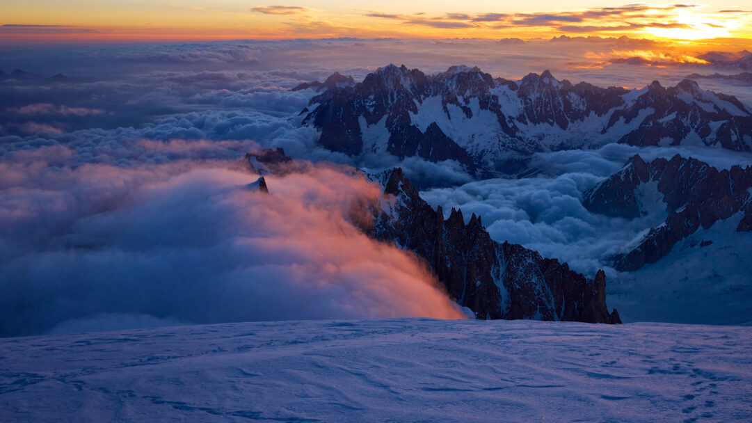 A breathtaking 4K wallpaper showcases the Mont Blanc Massif at sunset, rising majestically above a rolling sea of clouds near Chamonix, France. Warm oranges and vibrant pinks from the setting sun illuminate the distant horizon and touch the edges of the clouds, creating a dramatic contrast with the deep blue shadows across the snow-covered peaks and foreground.