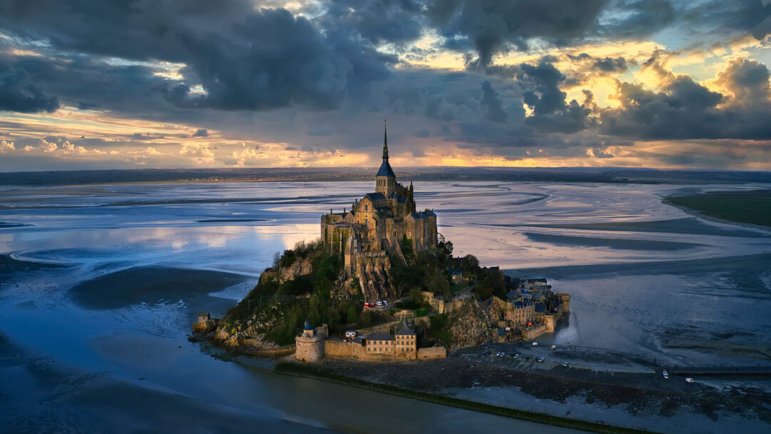 A captivating 4K wallpaper features Mont-Saint-Michel, a magnificent fortified abbey, standing proudly amidst expansive tidal flats in Normandy, France. The dramatic sunset casts golden light onto the towering structure and dark, brooding clouds, while deep blue hues reflect across the vast, wet tidal flats, evoking a majestic and ethereal mood.