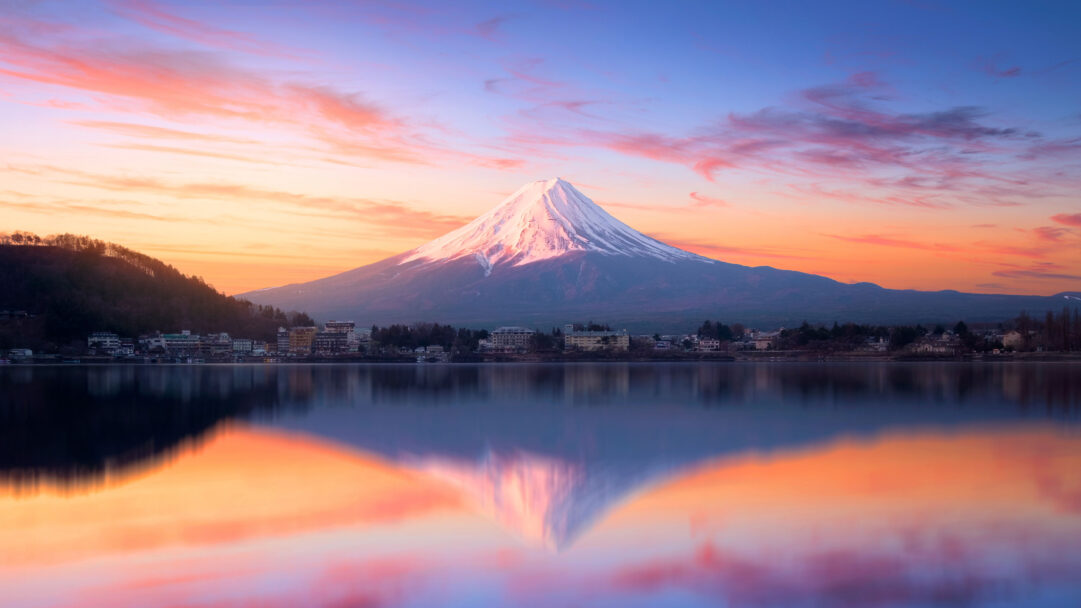 An awe-inspiring 4K wallpaper capturing Mount Fuji at sunrise over Lake Kawaguchi, with its snow-capped peak prominently overlooking a small town along the shore. The vibrant orange and pink clouds of dawn are perfectly mirrored in the tranquil waters, creating a symmetrical and ethereal landscape.