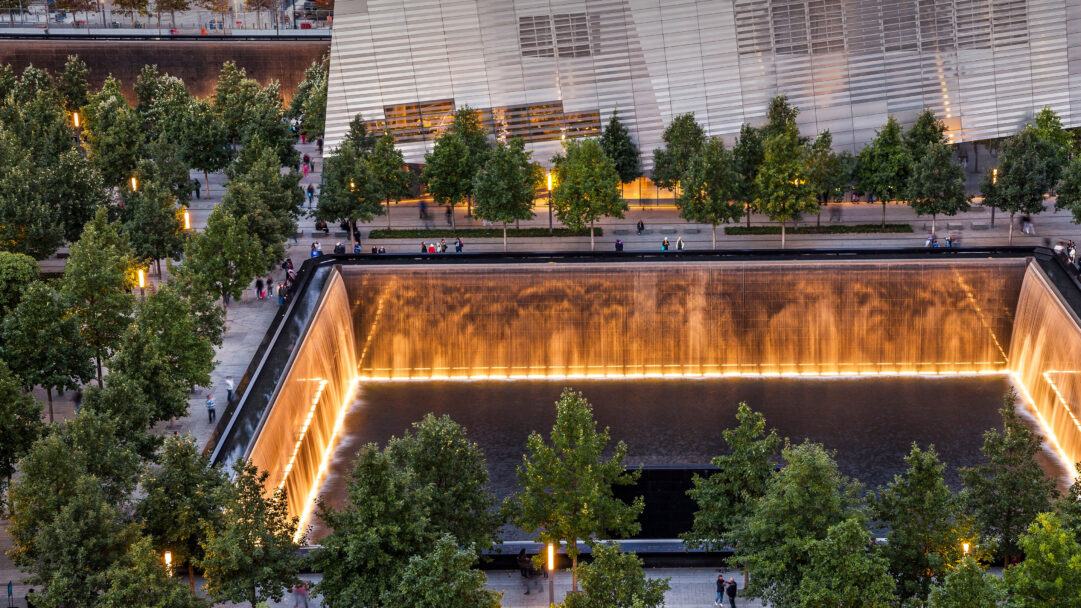 A solemn 4K wallpaper capturing the National September 11 Memorial's twin reflecting pools at dusk in New York City, surrounded by verdant trees. Golden light dramatically illuminates the cascading waterfalls, creating a profound and contemplative mood of remembrance against the twilight.