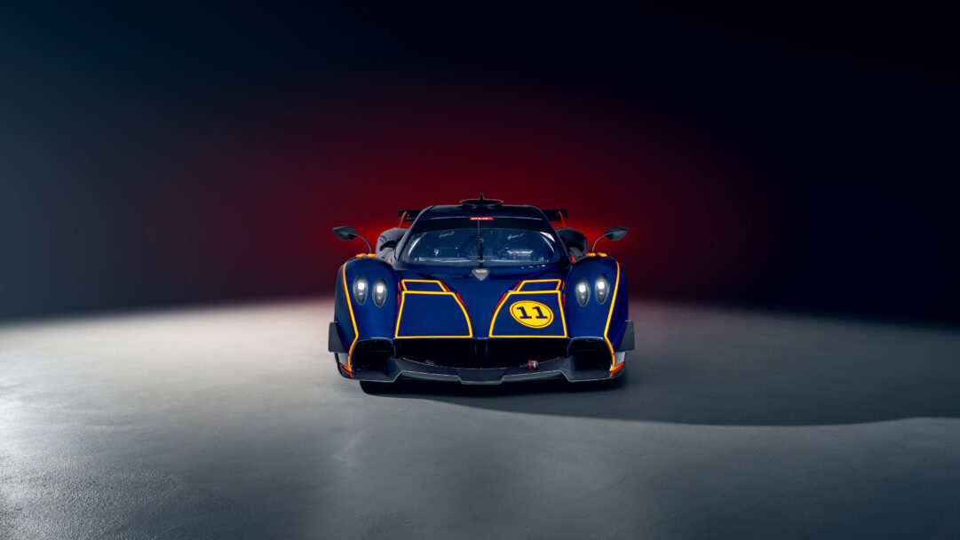 An exhilarating 4K wallpaper showcasing the iconic Pagani Huayra R supercar in a bold blue front view, positioned centrally within a dimly lit studio. Its intricate carbon fiber body, accented with vibrant yellow racing stripes and glowing headlights, creates a striking contrast against the dark, subtly red-tinged background and grey floor.