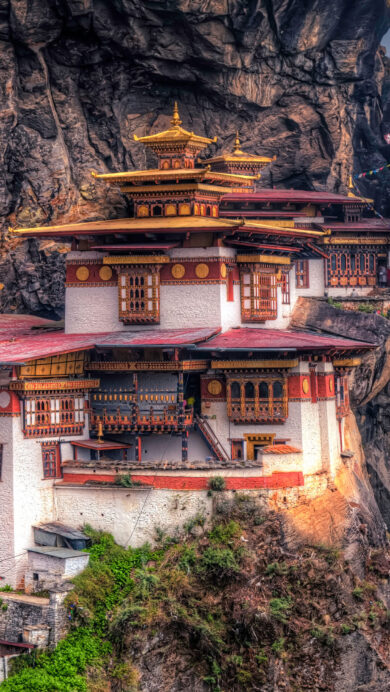 An iconic 4K wallpaper featuring the Paro Taktsang Buddhist Monastery, intricately built into a towering, rugged cliffside in Bhutan. The monastery's distinctive white walls, vibrant red roofs, and glittering golden spires beautifully contrast with the dark, textured rock and the misty, forested mountains in the distance, evoking a profound sense of spiritual majesty.