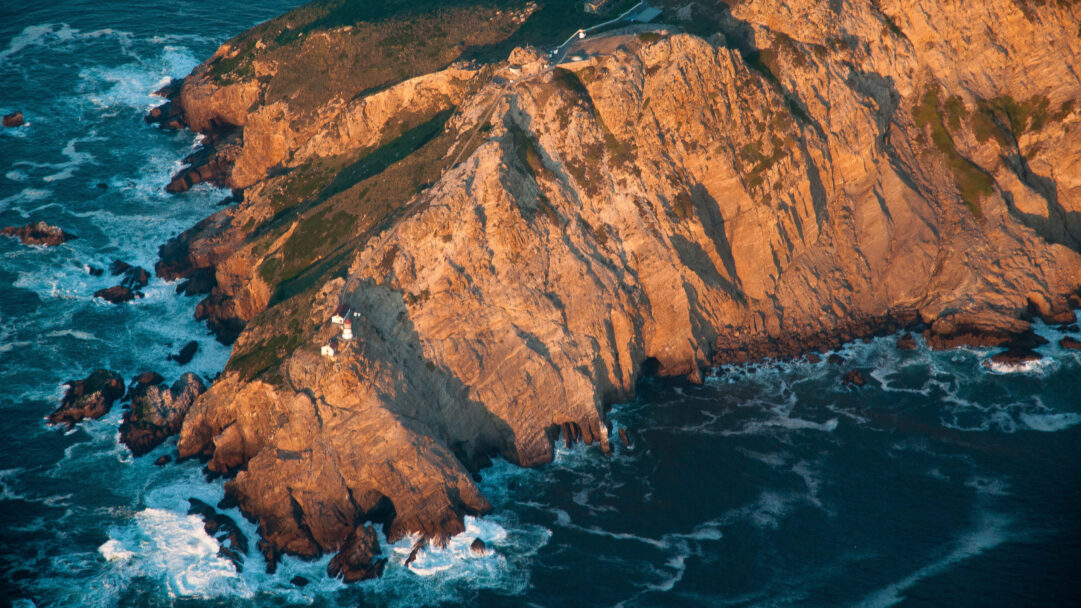 An awe-inspiring 4K wallpaper capturing the Point Reyes National Seashore Lighthouse perched dramatically on rugged, sunlit cliffs, seen from an aerial view overlooking the vast Pacific. The golden light of sunset illuminates the textured orange-brown cliffs, casting long shadows that sharply contrast with the deep teal-blue ocean churning with white foam below, evoking a sense of powerful natural beauty.