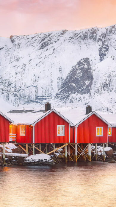 A breathtaking 4K wallpaper capturing red fishermen's cabins on the Reinefjorden shore, nestled at the base of majestic snowy mountains. The serene sunset casts a warm golden glow across the water and sky, beautifully contrasting with the cabins' vibrant red and the crisp white snow, creating a peaceful, inviting winter scene.