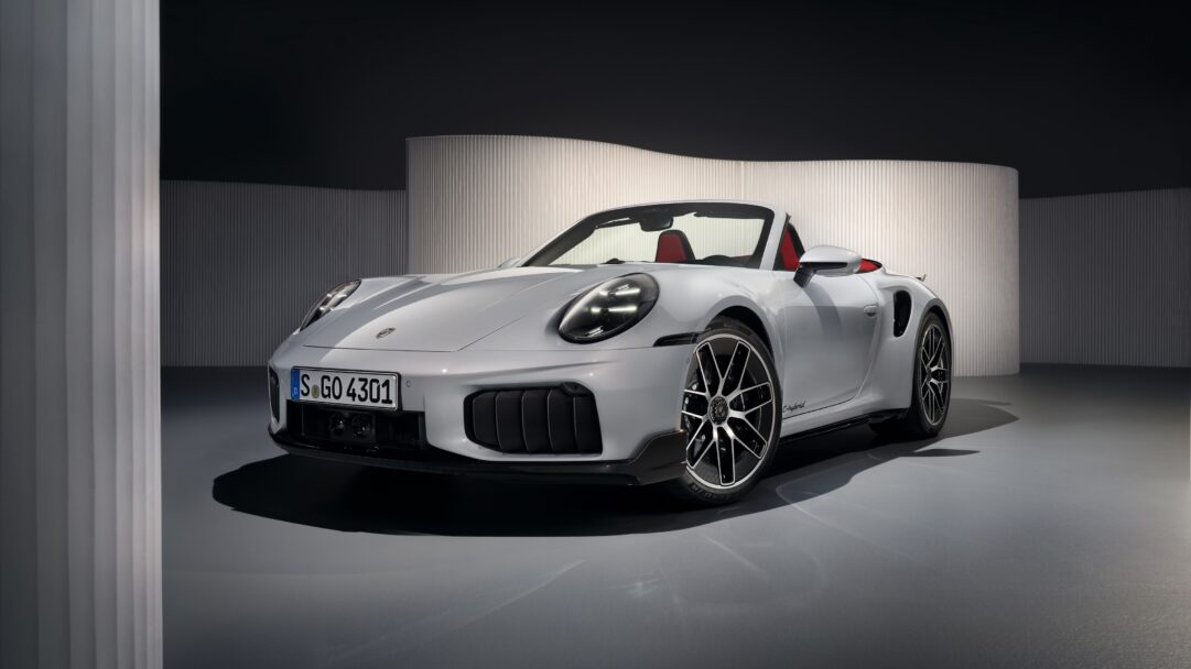 An exquisite 4K wallpaper presenting a sleek silver Porsche 911 Turbo S Cabriolet, prominently displayed within a sophisticated, minimalist studio environment featuring fluted architectural elements. Its metallic finish radiates under focused illumination, highlighting the sharp contours and the striking contrast of its red leather interior against the subdued grey and black background.