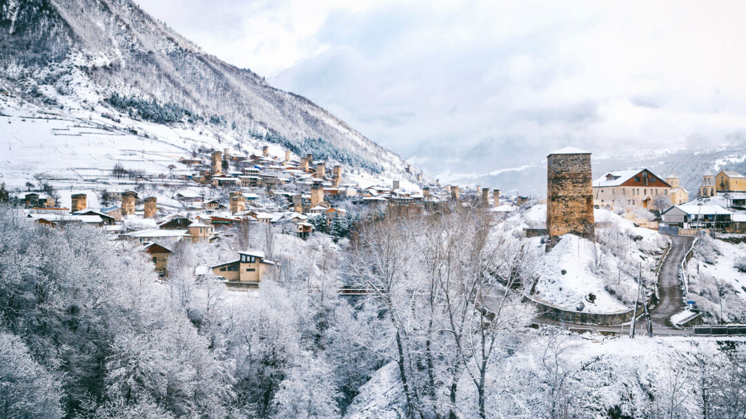 A majestic 4K wallpaper presents the snow-covered village of Mestia in Upper Svaneti, where numerous medieval stone towers rise strikingly from the winter landscape. The pristine white snow blankets every surface, from the dense forest in the foreground to the distant mountain peaks, creating a serene and timeless winter panorama under a soft, cloudy sky.