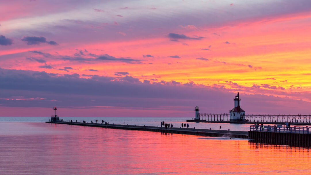 A breathtaking 4K wallpaper showcasing the St. Joseph North Pier with its Inner and Outer Lights extending into the tranquil water, where people stroll along the long pier. The dramatic pink and orange sunset paints the sky and reflects intensely on the water, creating a serene and captivating evening atmosphere.
