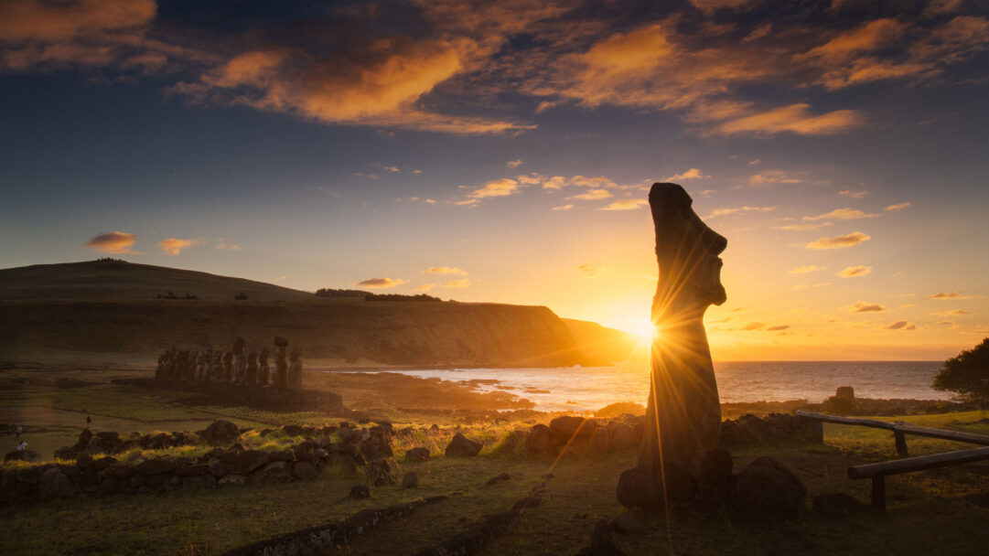 An awe-inspiring 4K wallpaper unveils the majestic sunrise at Ahu Tongariki, Rapa Nui National Park, with an iconic Moai statue silhouetted against the horizon and other colossal figures lined in the distance along the rugged coastline. The brilliant golden sun bursts from directly behind the foreground Moai, creating dramatic rays that illuminate the orange-streaked clouds and infuse the ancient landscape with a sense of profound spiritual awakening.