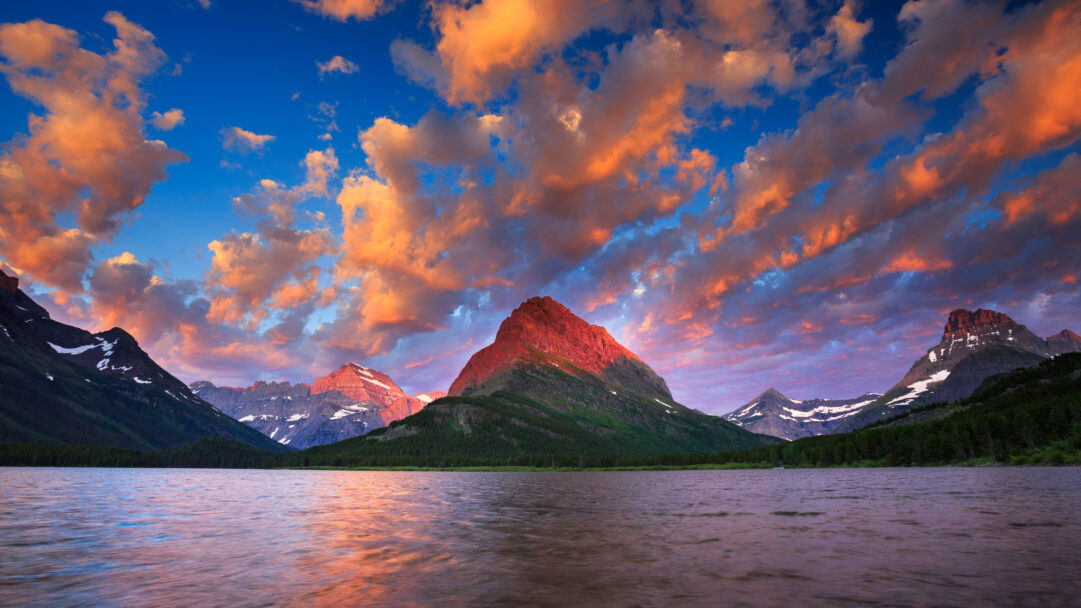 A breathtaking 4K wallpaper featuring a dramatic sunrise over Swiftcurrent Lake, nestled among majestic mountains. Fiery orange and red clouds illuminate the sky and reflect vividly on the lake's rippling surface, bathing the prominent mountain peak in a warm, ethereal glow.