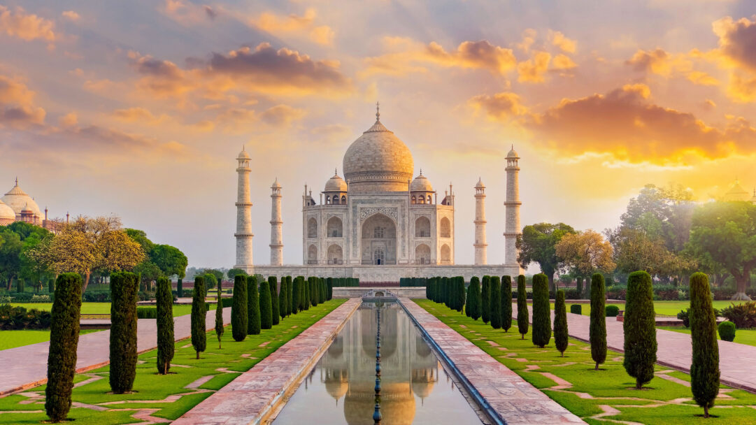 A breathtaking 4K wallpaper showcases the majestic Taj Mahal in Agra, India, presiding over symmetrical gardens and a long reflecting pool at sunset. The white marble structure glows subtly under a sky painted with warm oranges and soft purples, perfectly mirrored in the still water below, enhancing its serene and timeless beauty.