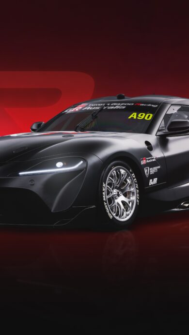 A dynamic 4K wallpaper presents a sleek, matte black Toyota GR Supra Racing Supercar, adorned with white and red racing decals, parked on a reflective surface against a rich, gradient red background. Its piercing LED headlights and metallic wheels shimmer, contrasting with the car's matte finish and the deep red hues of the backdrop, creating an intense, high-performance atmosphere.