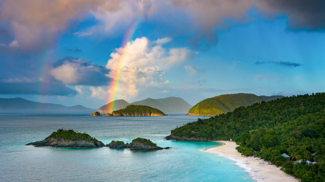 A breathtaking 4K wallpaper captures Trunk Bay in St. John, Virgin Islands National Park, with its pristine white sand beach curving along turquoise waters and small green islets. A vivid rainbow arches gracefully across the dramatic sky, casting brilliant hues over the lush green hills and sun-drenched sea, creating a serene and magical tropical vista.