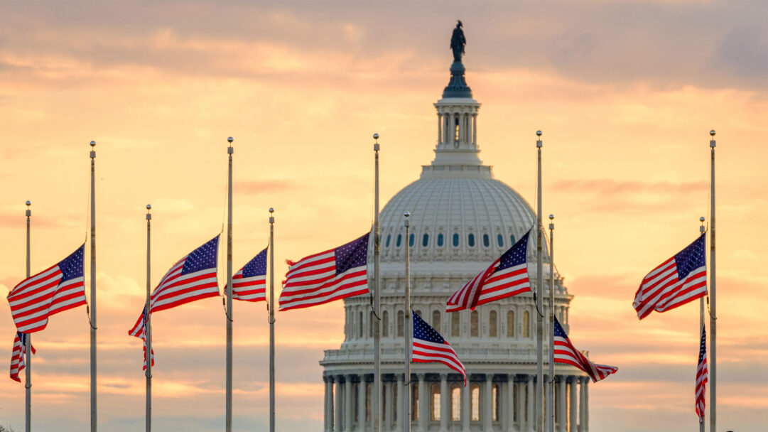 An iconic 4K wallpaper capturing the US Capitol Dome framed by numerous American flags. The setting sun bathes the scene in warm golden hues, highlighting the flags waving proudly against the twilight sky.