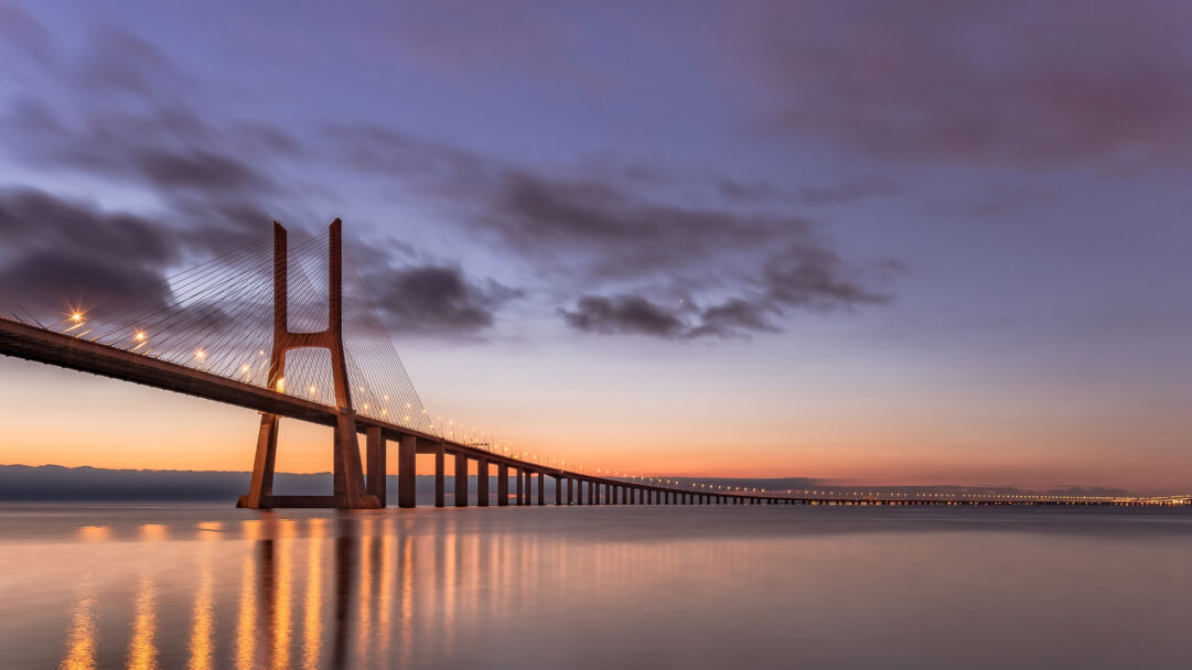 A breathtaking 4K wallpaper showcasing the immense Vasco da Gama Bridge spanning across the waters near Lisbon, Portugal, bathed in the soft hues of twilight. Its illuminated streetlights cast long, golden reflections upon the tranquil water, creating a serene and expansive panorama against the deepening purple sky.