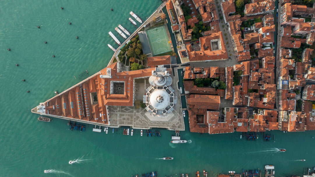 A breathtaking 4K wallpaper showcasing an aerial view of Venice's Grand Canal and the iconic Basilica Santa Maria Della Salute. The vibrant turquoise water of the canal, traversed by numerous boats, contrasts beautifully with the dense labyrinth of terracotta rooftops that surround the majestic white dome of the basilica, creating a stunning visual tapestry.