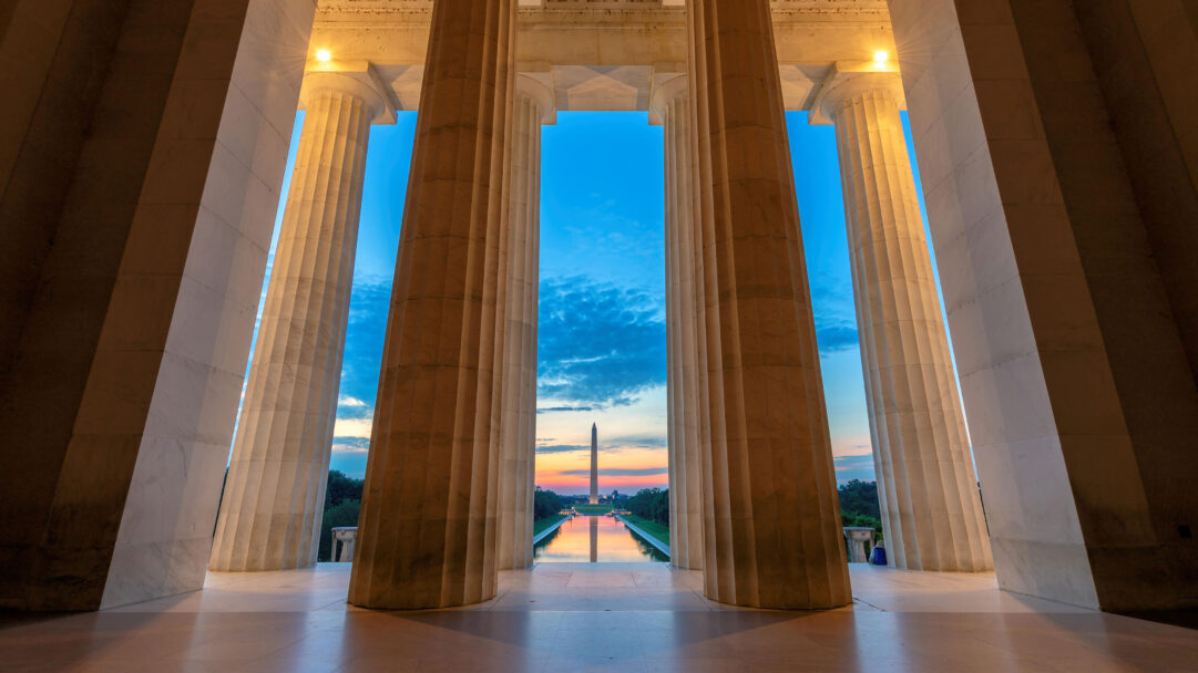An iconic 4K wallpaper capturing the Washington Monument, framed dramatically by the towering pillars of the Lincoln Memorial, extends across the Reflecting Pool at sunset. The monument rises silhouetted against a brilliant sky transitioning from deep blue to soft pastels of orange and pink, beautifully reflected in the calm waters below, creating a powerful sense of national grandeur.