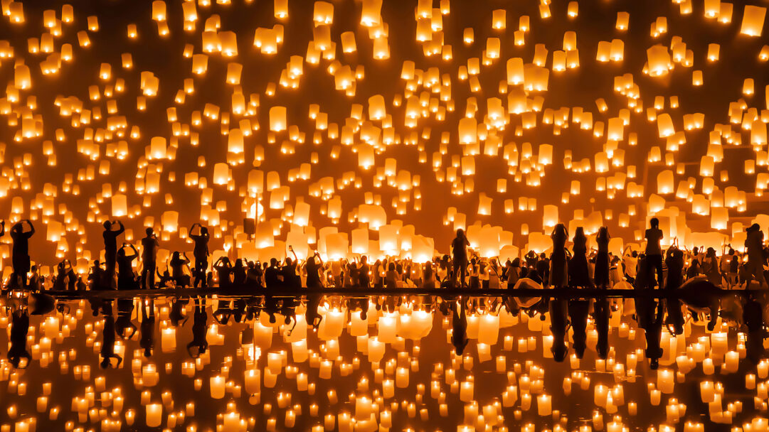 An enchanting 4K wallpaper transports you to the Yi Peng Lantern Festival in Chiang Mai, Thailand, where countless sky lanterns ascend into the dark night. This stunning spectacle is magnificently doubled by a perfect water reflection, illuminating the silhouetted crowds below in a vibrant, golden glow.