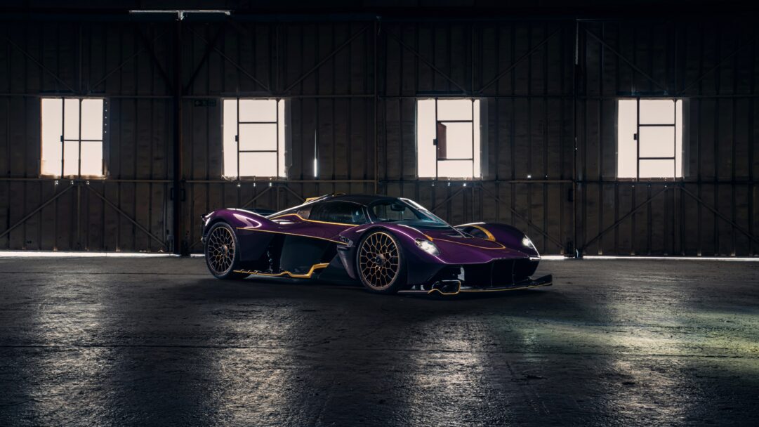 An electrifying 4K wallpaper showcases a vibrant purple and gold Aston Martin Valkyrie hypercar parked centrally within a vast, dimly lit hangar. Its sleek bodywork, highlighted by subtle light filtering through the large windows behind, reflects brilliant gold accents that amplify its luxurious and powerful presence.
